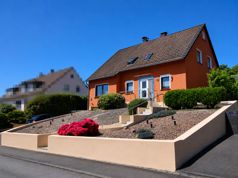 A two-story orange house with a brown roof, featuring a terraced front garden with red flowers and a concrete staircase.