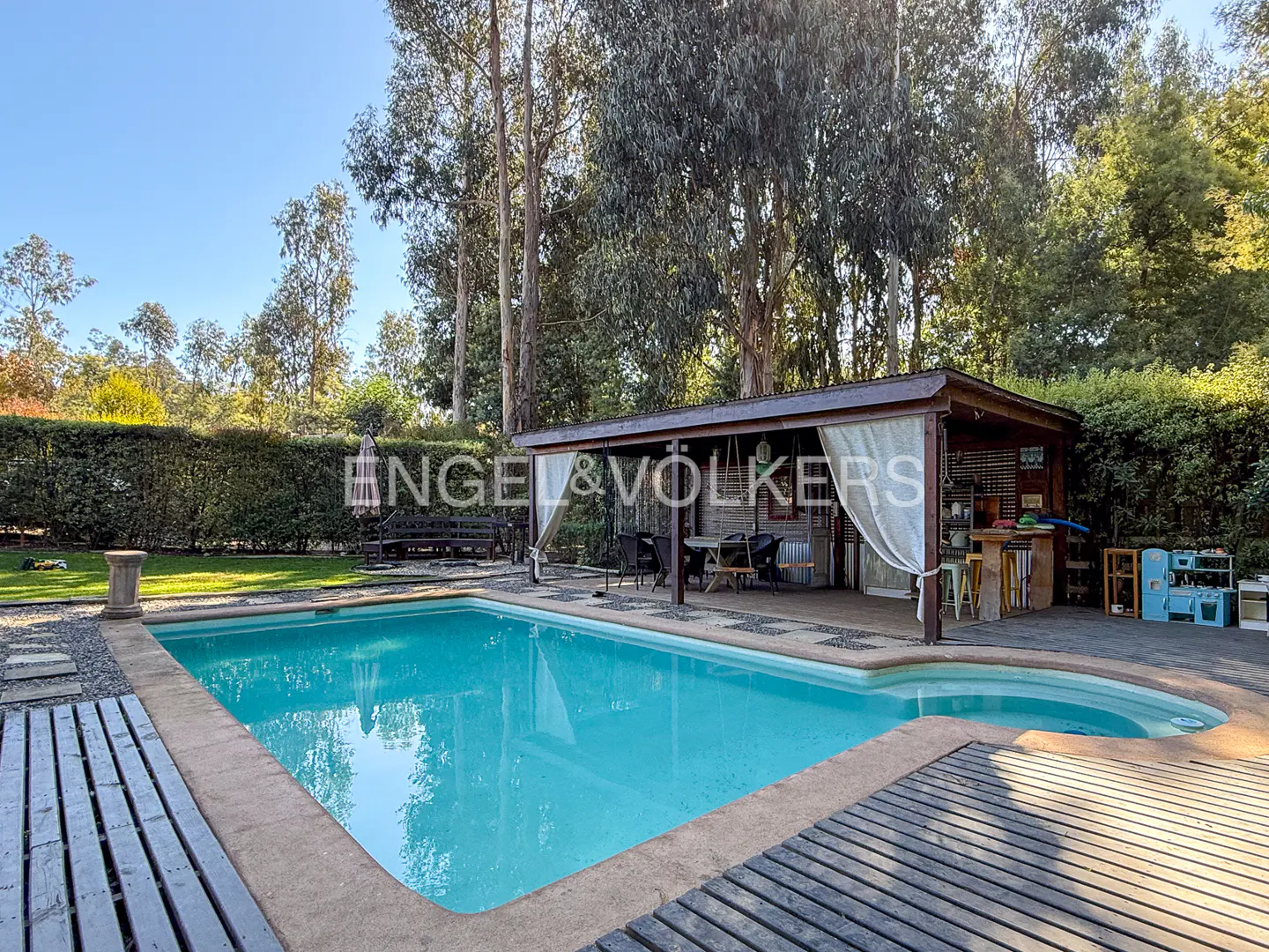 A backyard with a blue swimming pool, a wooden deck, and a cabana with white curtains. Tall trees and green bushes surround the area.
