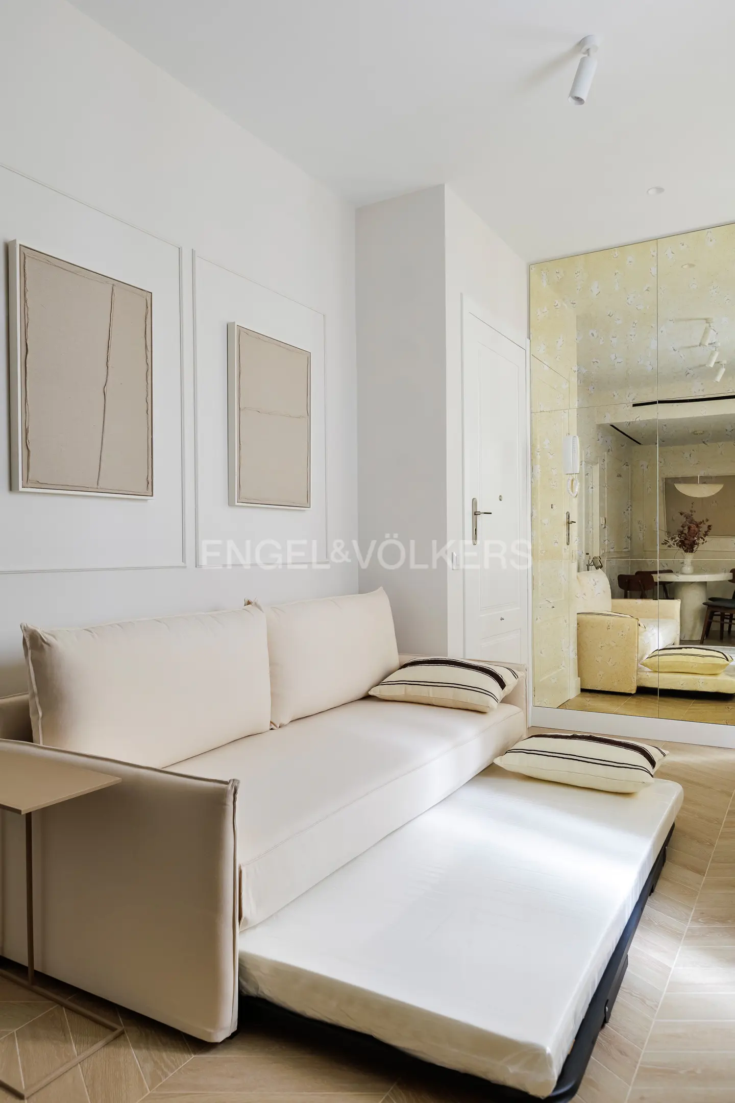 Living room with a pull-out sofa bed, beige walls, and two framed abstract art pieces. A mirrored wall reflects the dining area.