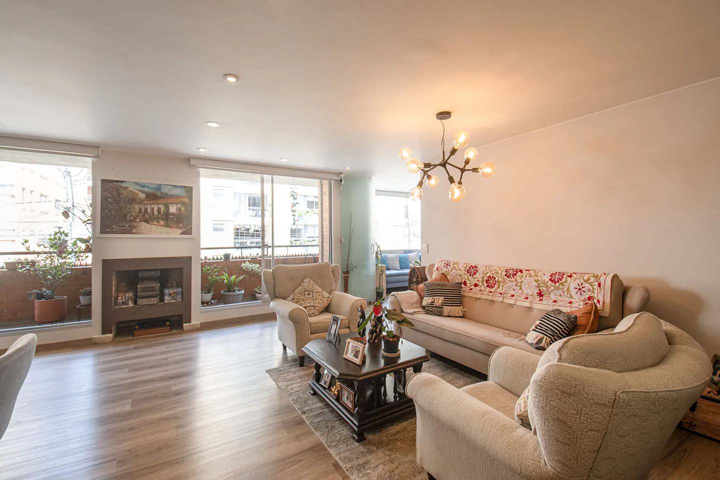 Bright living room with hardwood floors, beige sofa and armchairs, a dark wood coffee table, and a modern chandelier. Large windows offer a city view.