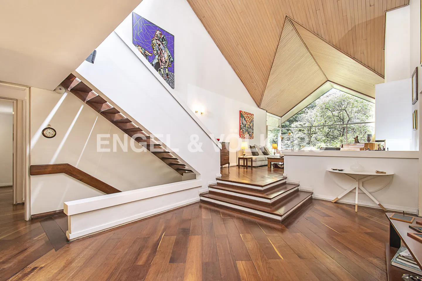 Interior view of a modern home with wood floors, stairs, and a high, angled wood ceiling. A large window overlooks lush greenery. Artwork adorns the white walls.