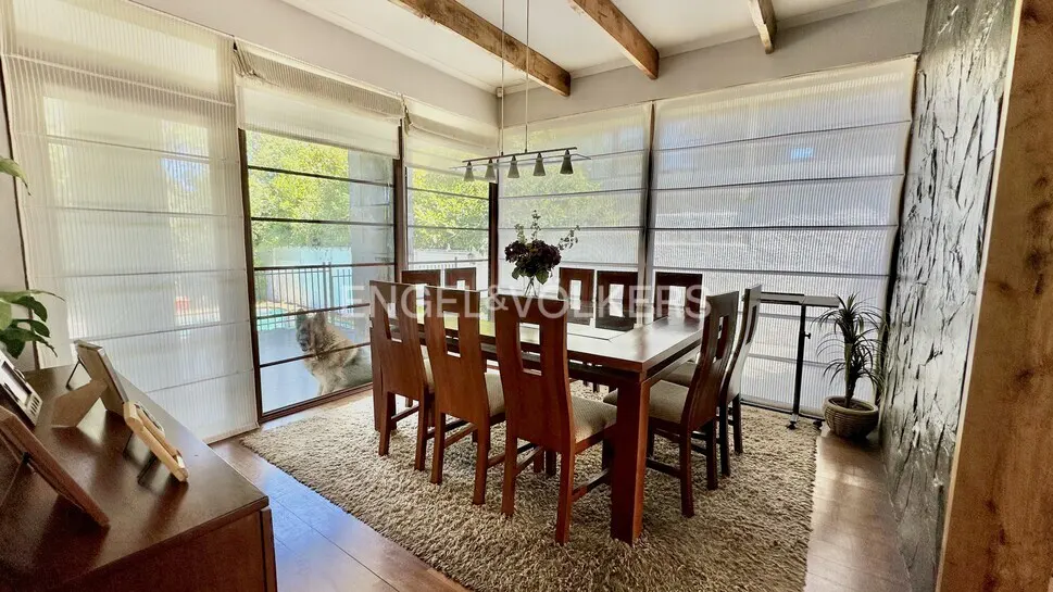 A dining room with a long wooden table and chairs on a beige rug, with large windows and a dog outside.