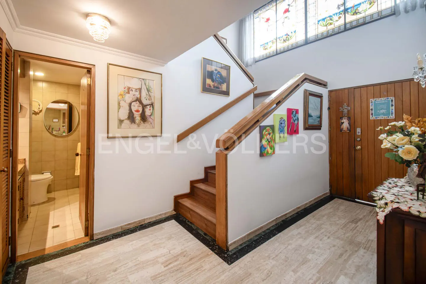 A home's foyer with a wooden staircase, artwork on the walls, and a bathroom visible through an open door.
