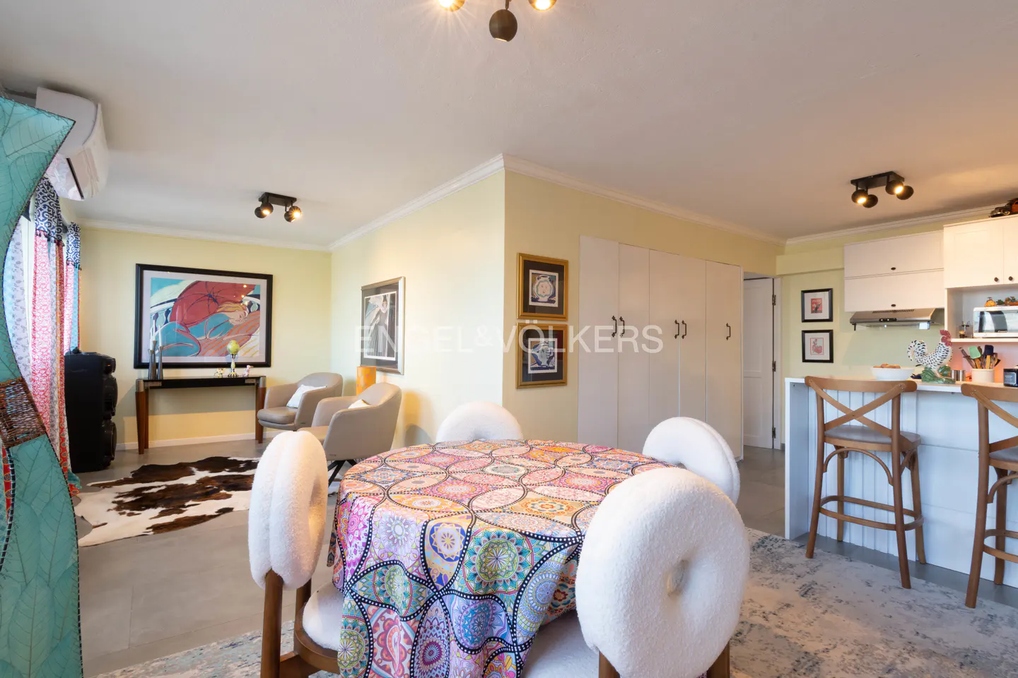 A bright, open-concept living space with a round dining table covered in a colorful patterned tablecloth. White chairs surround the table. A kitchen and seating area are visible.