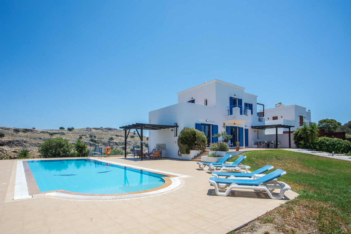 White villa with blue shutters, a swimming pool, and lounge chairs on a sunny patio against a clear blue sky. [AI-generated text]