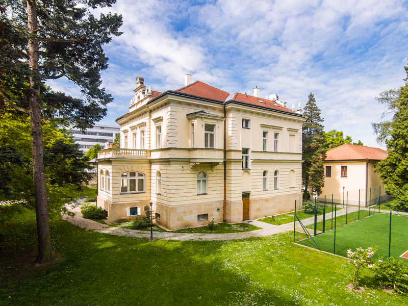 A grand, cream-colored historic villa with a red tile roof sits in a lush green garden next to a small sports court.