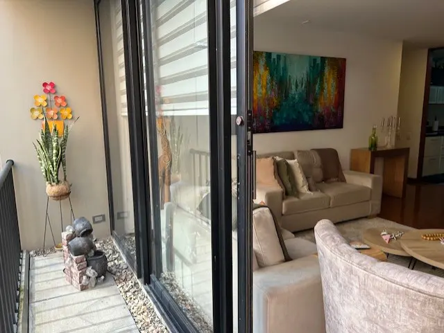 A balcony with a snake plant and fountain overlooks a modern living room with a beige sofa and colorful abstract art.