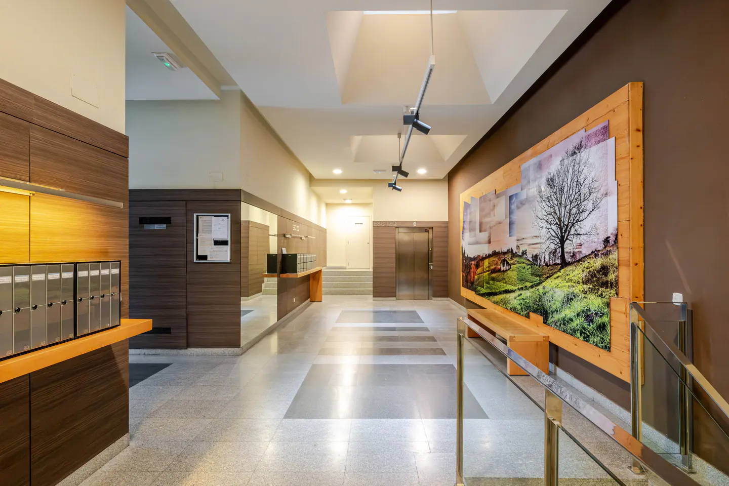 Apartment lobby with mailboxes, elevator, and large landscape art on a brown wall.