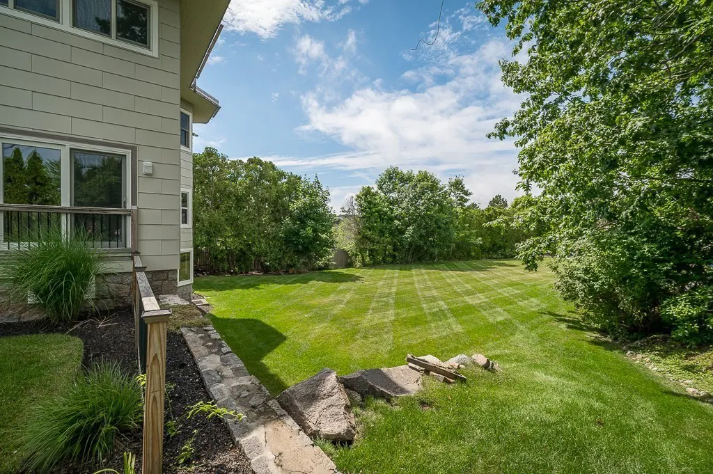 A view of a large, manicured backyard with a stone retaining wall, lush green grass, and trees under a blue sky.