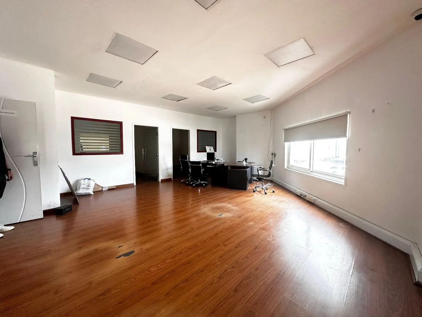 Empty office space with wood floors, white walls, and a desk with chairs. Natural light from a window with a roller blind.