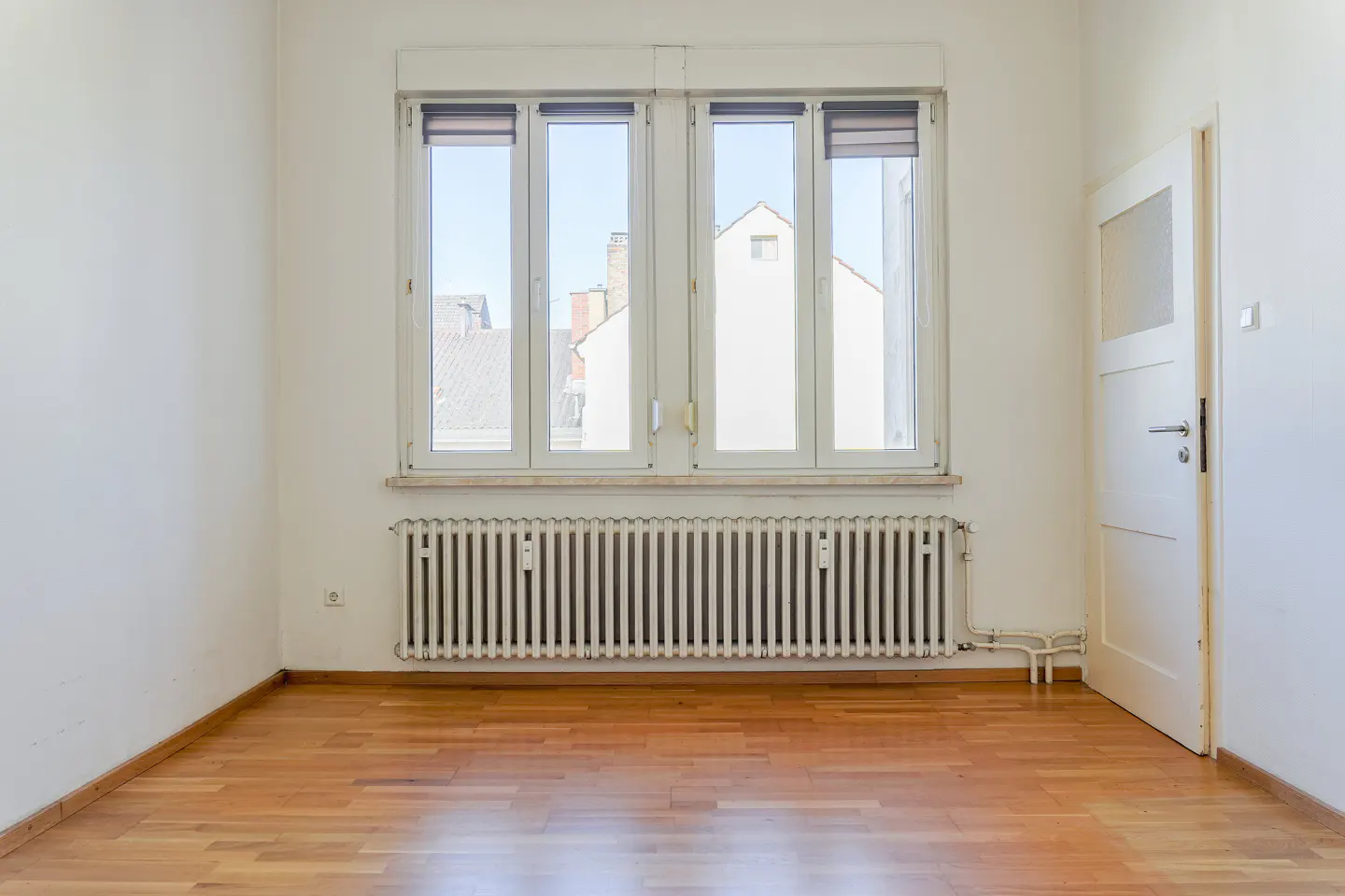 Bright, empty room with wood floors, white walls, radiator under a window, and a partially open white door.