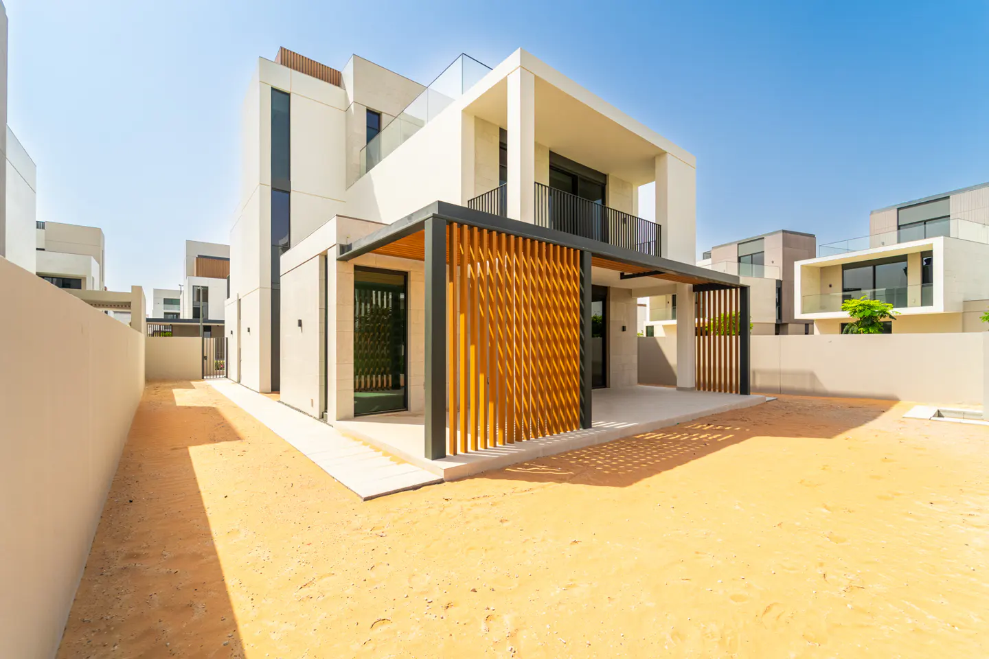 Modern two-story white house with a wood slat pergola and a sandy yard under a clear blue sky.