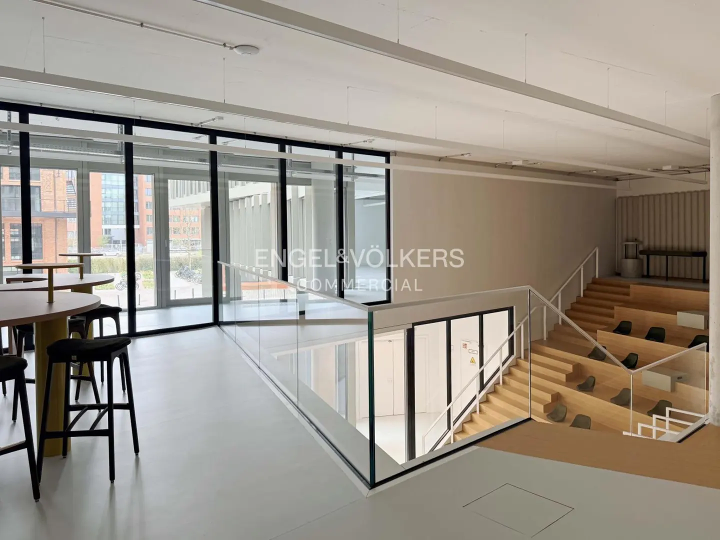 Bright, modern office space with tables, chairs, and tiered seating. Engel & Völkers Commercial logo visible.