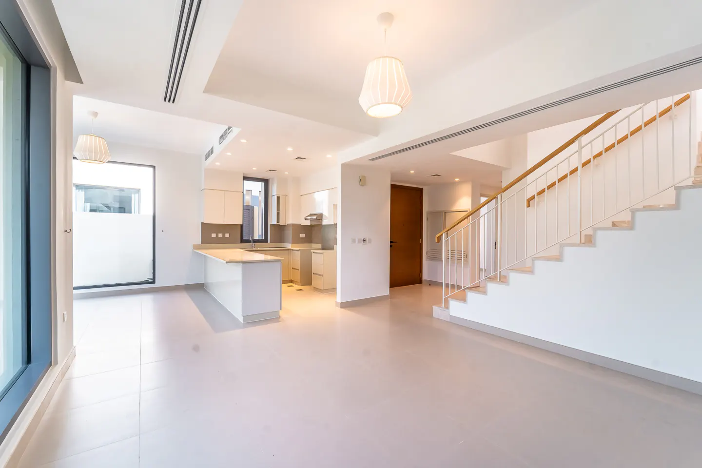 Bright, open-plan living space with white walls, tile floor, kitchen island, and staircase with wooden handrail.