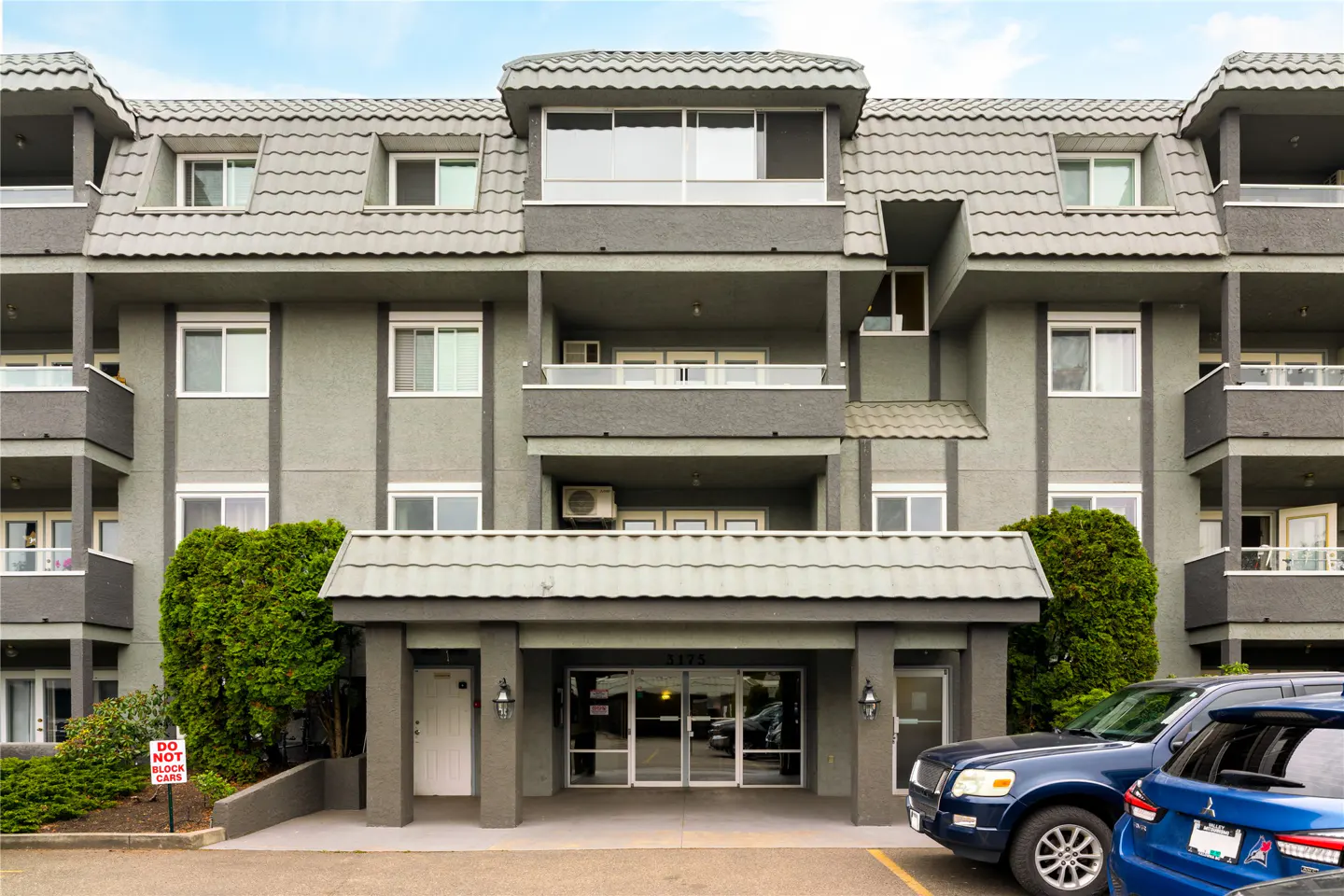 Exterior of a gray apartment building with balconies, a tiled roof, and a covered entrance. Cars are parked in front.