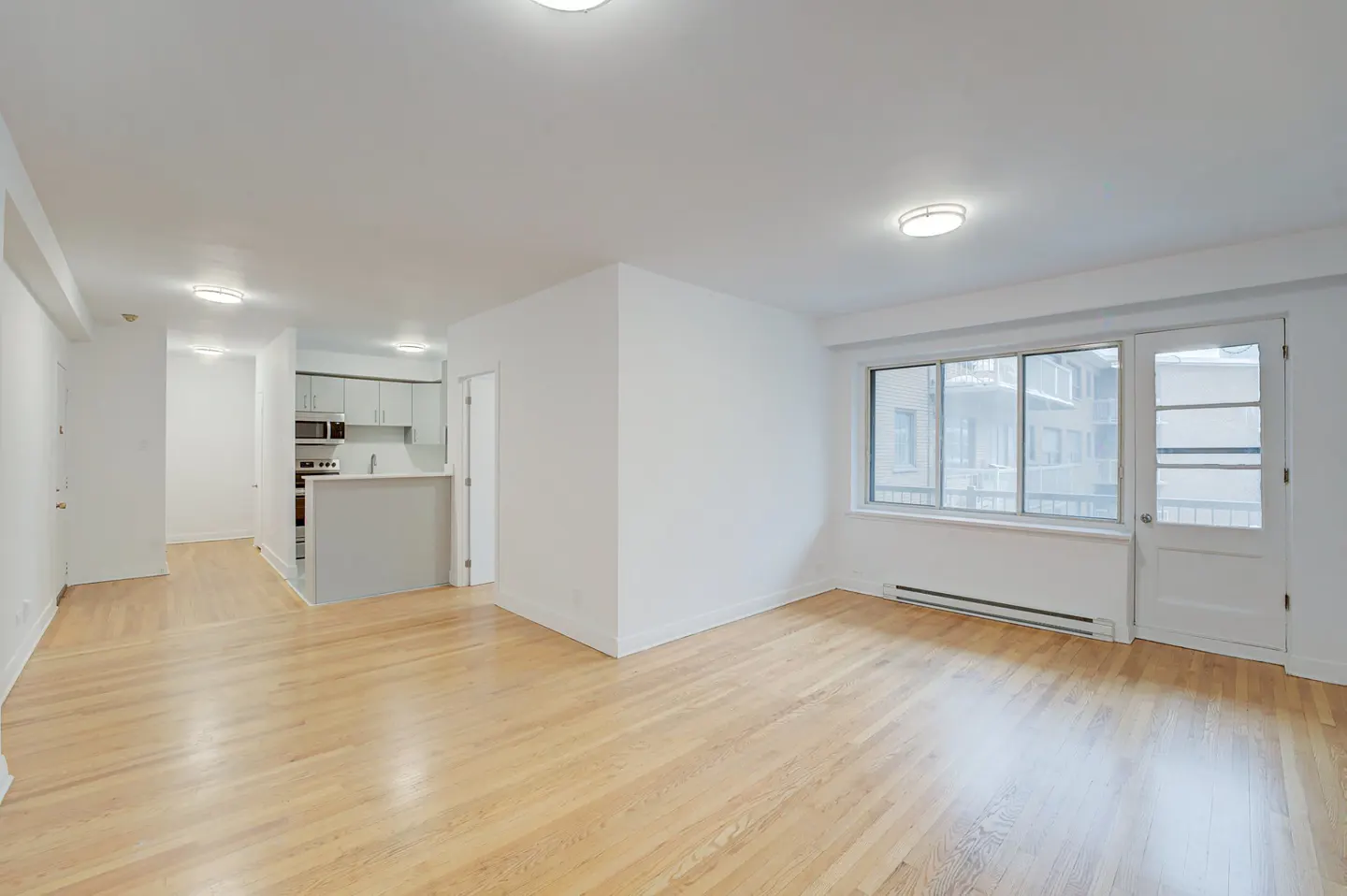 Bright, empty apartment with hardwood floors, white walls, and recessed lighting. Kitchen visible in the background. Large window and door to balcony.