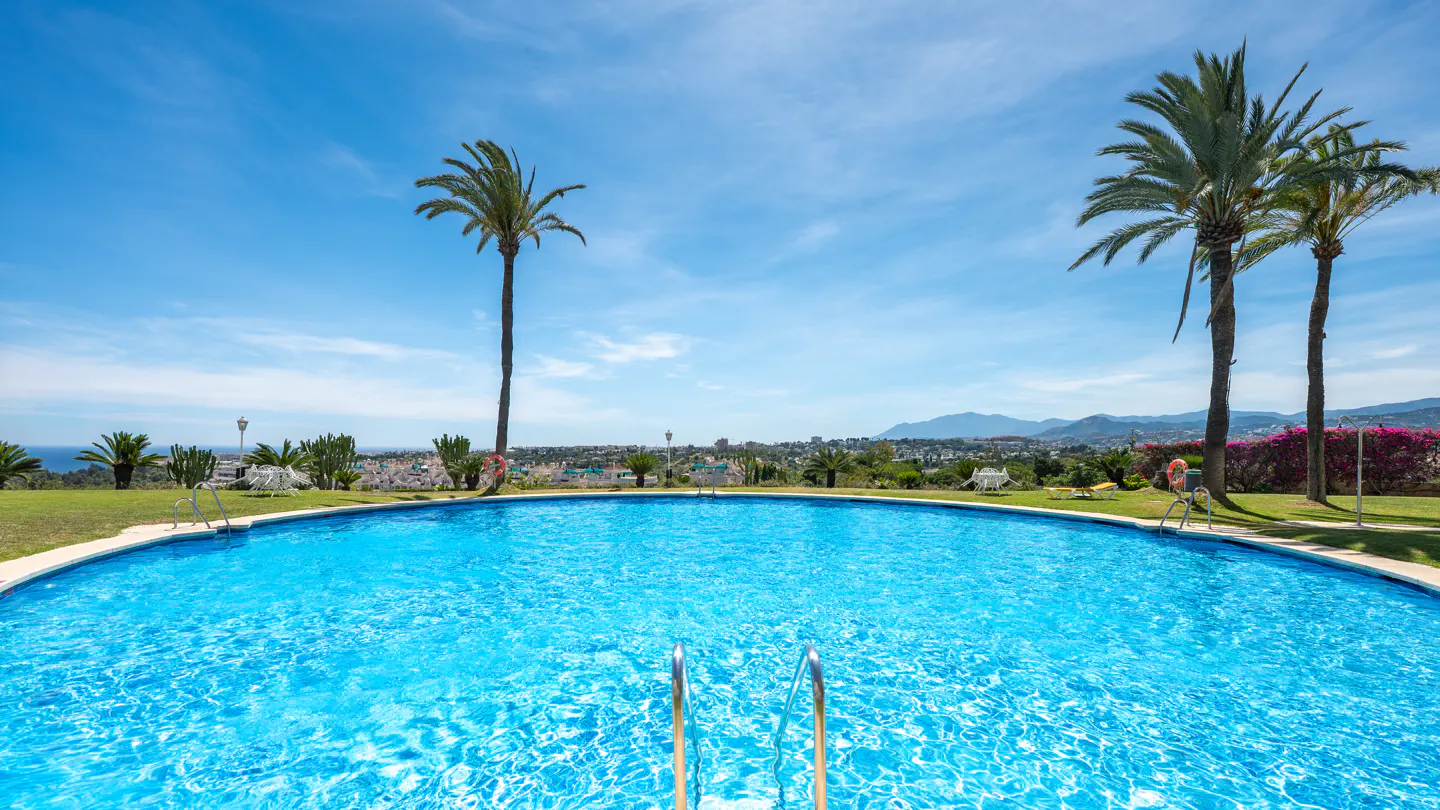 A bright blue circular swimming pool with metal ladders, surrounded by palm trees and a scenic coastal town view.