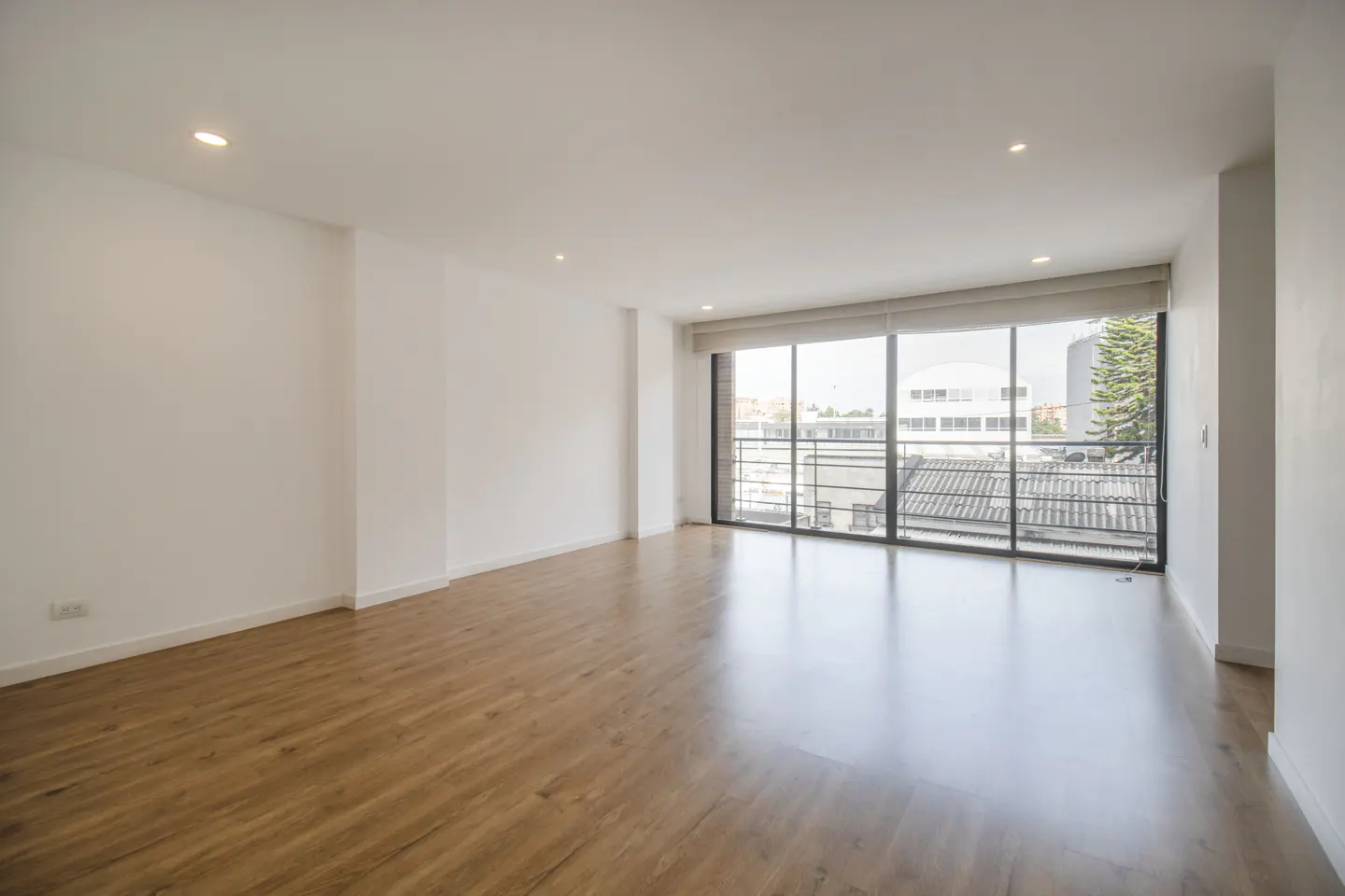 Bright, empty room with white walls, wood floors, and a large window overlooking a city.