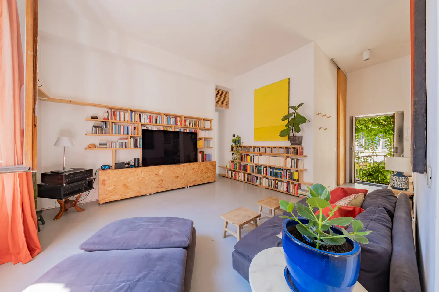 Modern living room with a grey sofa, wooden shelving units, a large TV, a bright yellow wall art, and a blue potted plant.