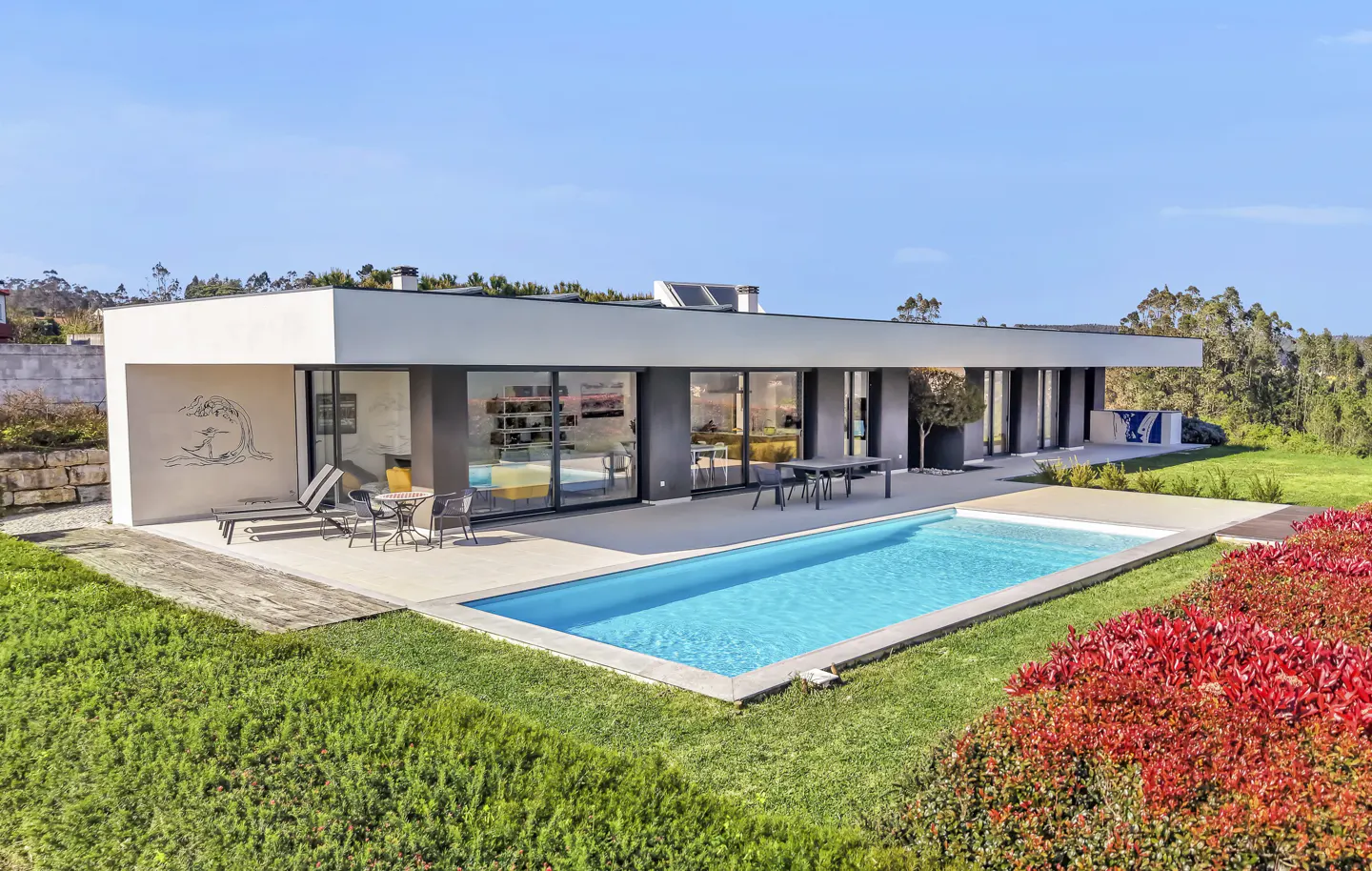 Modern home with a pool. The house has large glass windows and a flat roof. There is a patio with lounge chairs and a table.
