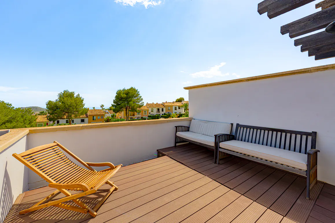 Outdoor patio with wooden deck, white walls, and blue sky. A wooden lounge chair and benches with cushions offer seating. Houses and trees are visible in the background.