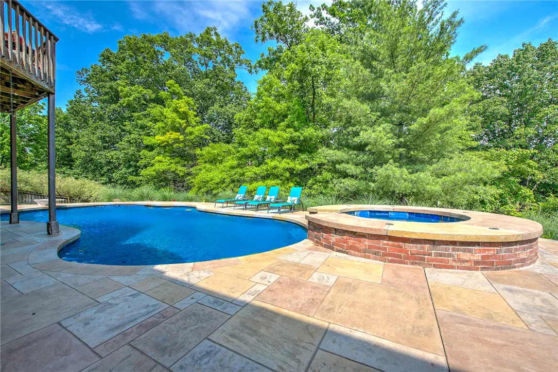 Backyard pool with blue water, stone patio, and brick hot tub. Four blue lounge chairs sit poolside, with green trees in the background.