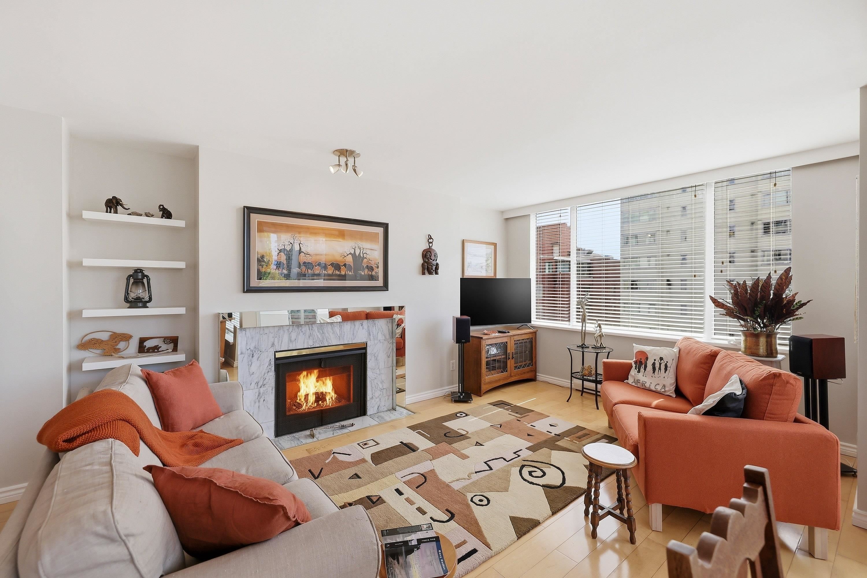 Living room with a fireplace, orange sofa, and beige sofa. Shelves with decor, a TV, and a patterned rug are also visible.