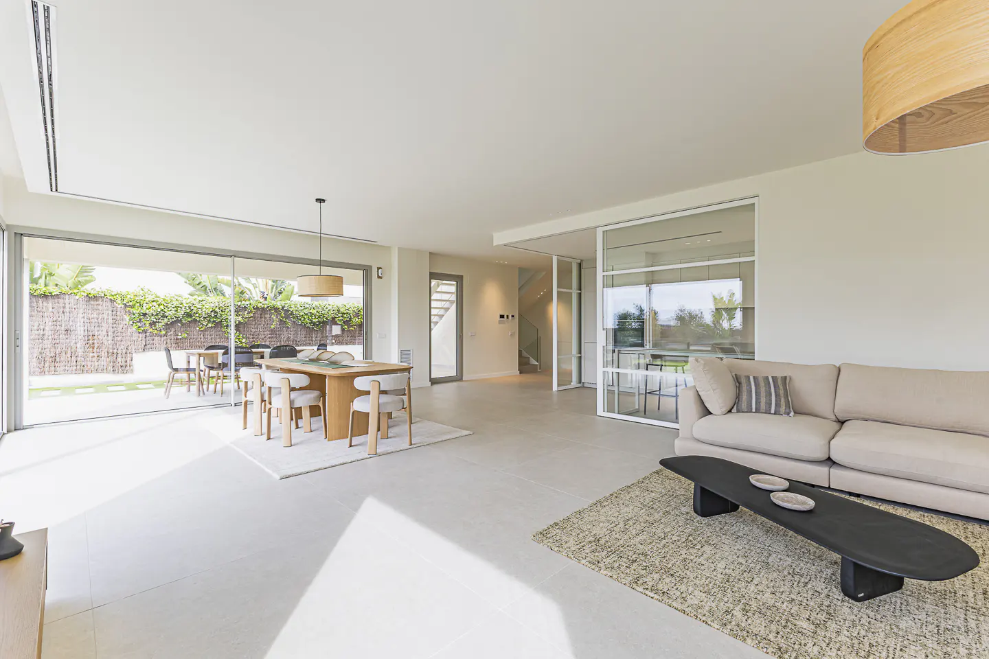 Bright, open-plan living space with a beige sofa, black coffee table, and a wooden dining table with six chairs. Sliding glass doors lead to a patio.