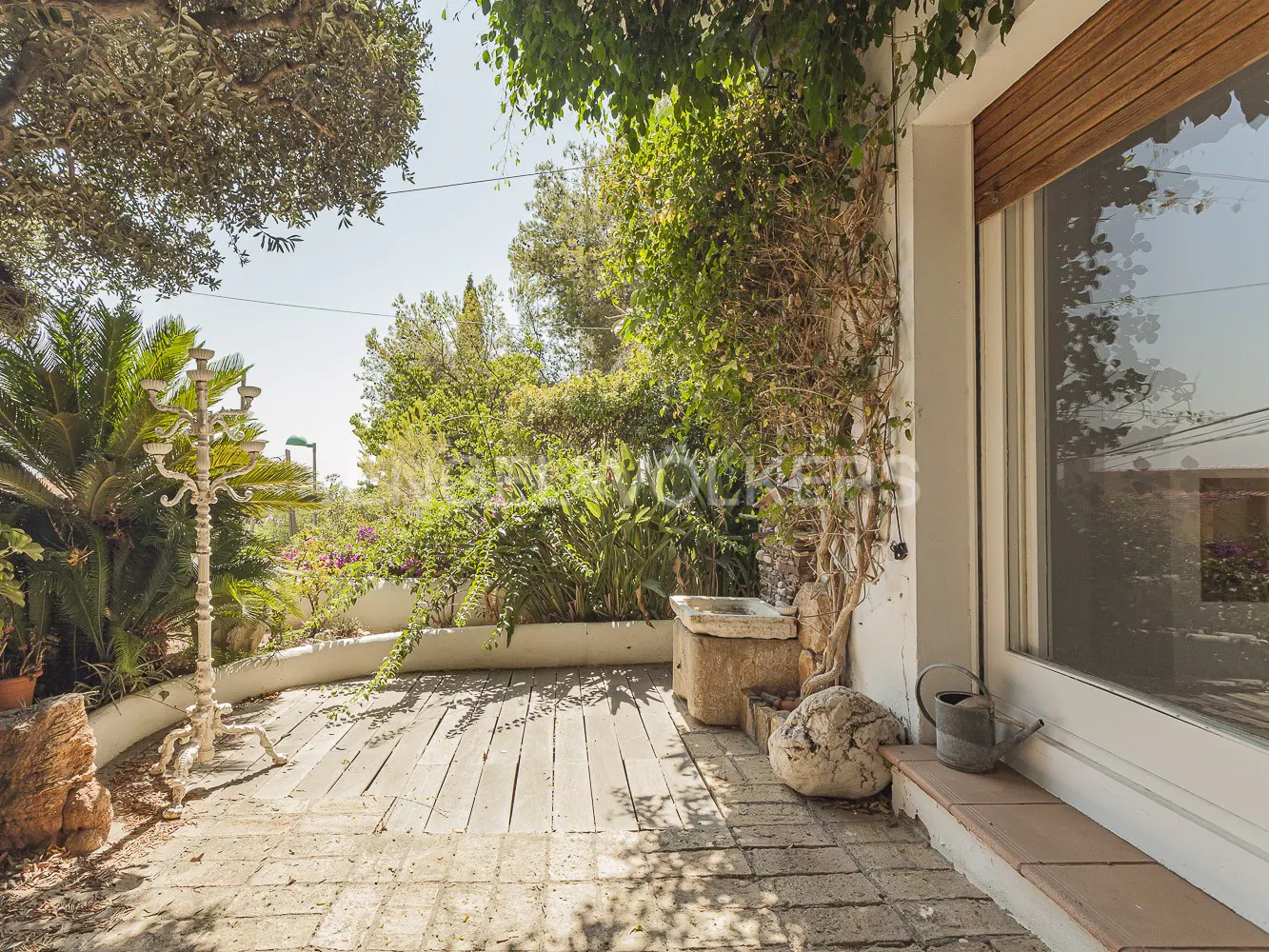 Outdoor patio with stone flooring, lush greenery, and a white building with a large window. A vintage candelabra and watering can add charm.