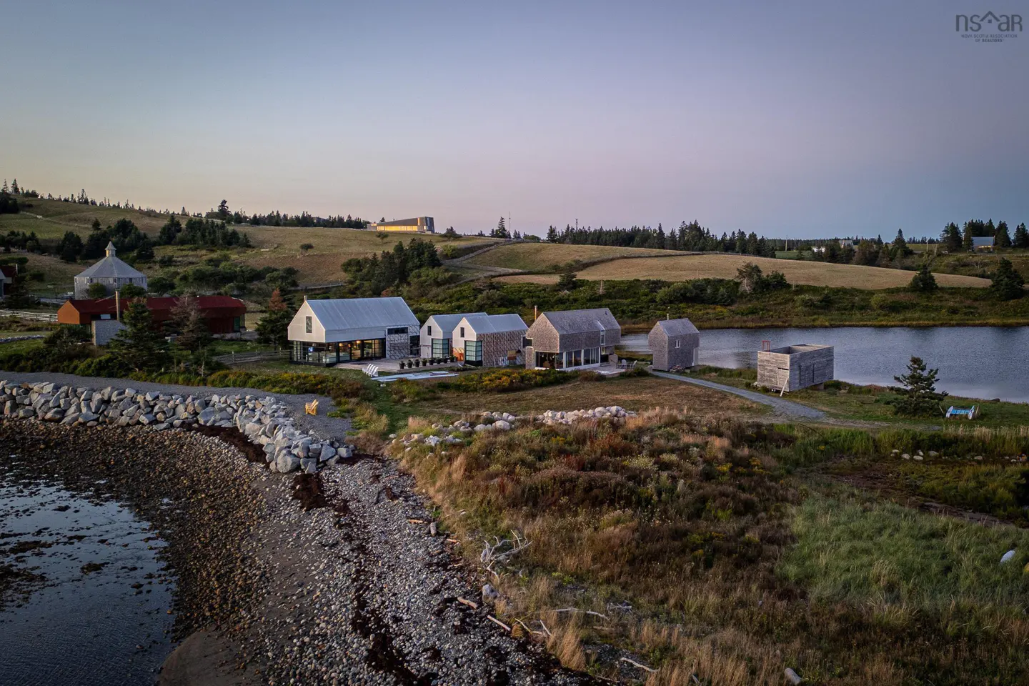 Aerial view of modern waterfront homes with metal roofs, near a rocky beach and calm water, set against a rural landscape.