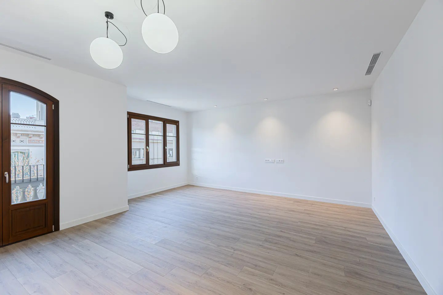 Bright, empty room with light wood floors, white walls, and dark wood trim on the arched door and window. Two globe lights hang from the ceiling.