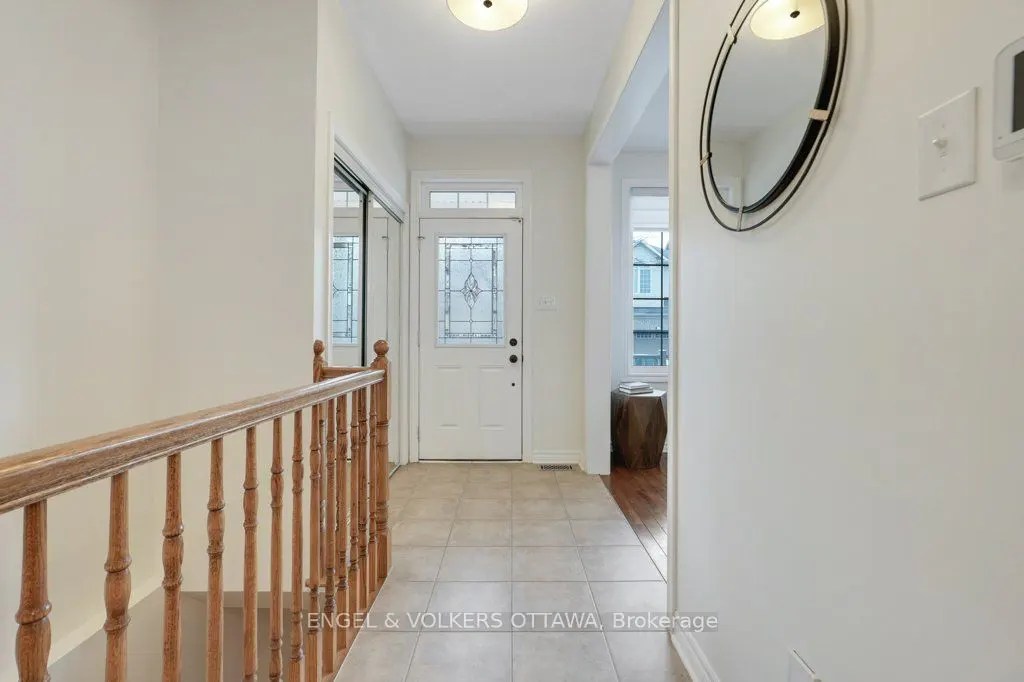 Bright foyer with white walls, tile floor, and wood railing. A white front door with glass detail is centered. An oval mirror hangs on the right wall.