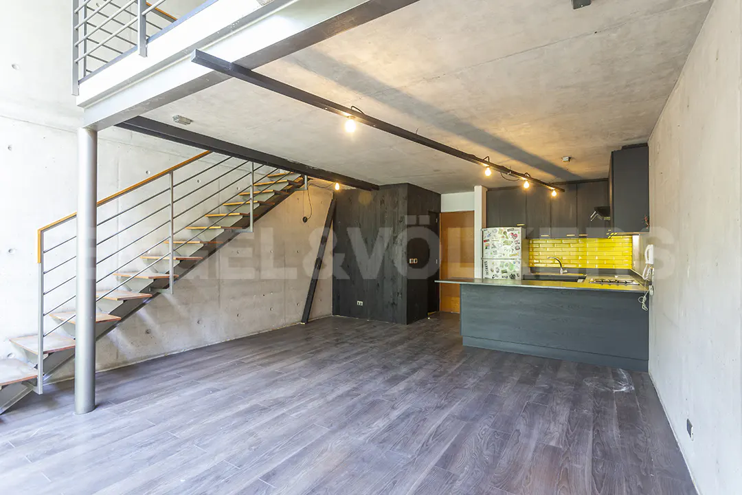 Open-concept loft apartment with gray wood floors, concrete walls, and a modern kitchen with yellow backsplash. Stairs lead to a second level.