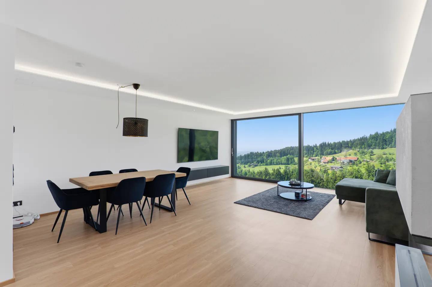 Bright, modern living room with wood floors, dining table with blue chairs, and a large window overlooking a green, hilly landscape.