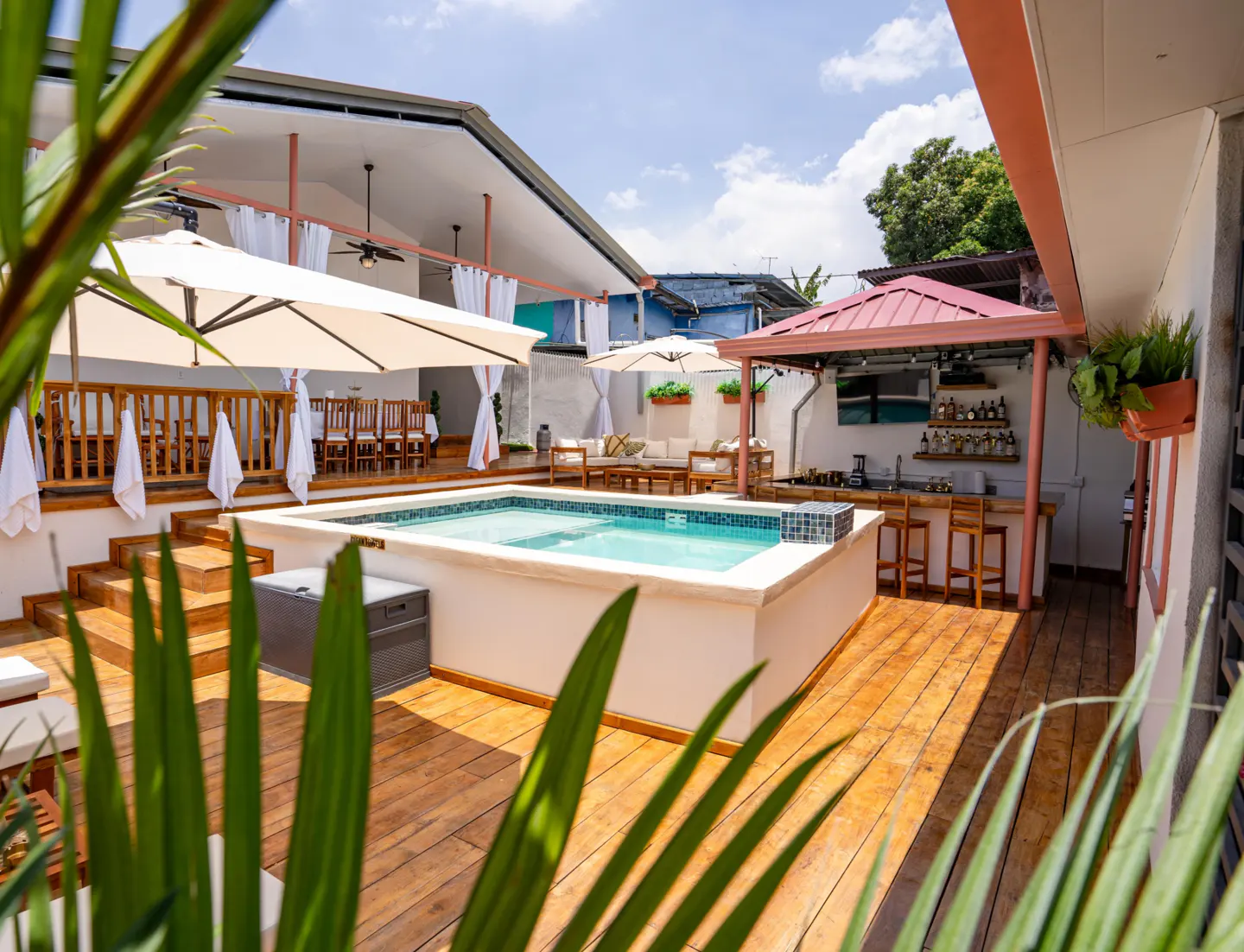 Outdoor living space with a pool, wooden deck, dining area, lounge, and bar under a red-roofed gazebo on a sunny day.
