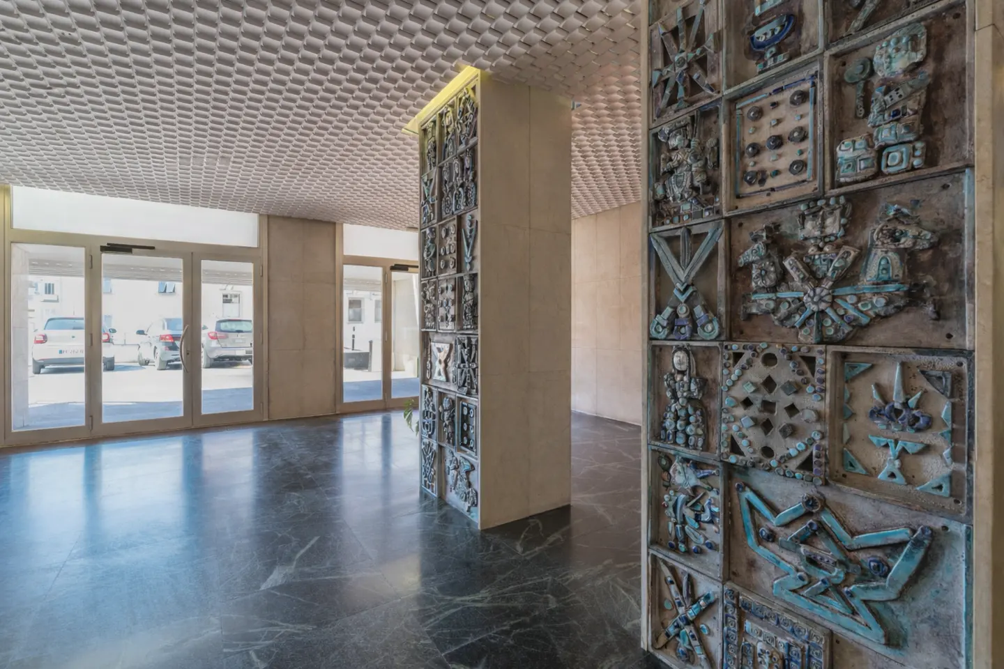 Lobby with marble floor, textured ceiling, and decorative columns with blue-green tile art. Glass doors lead outside.