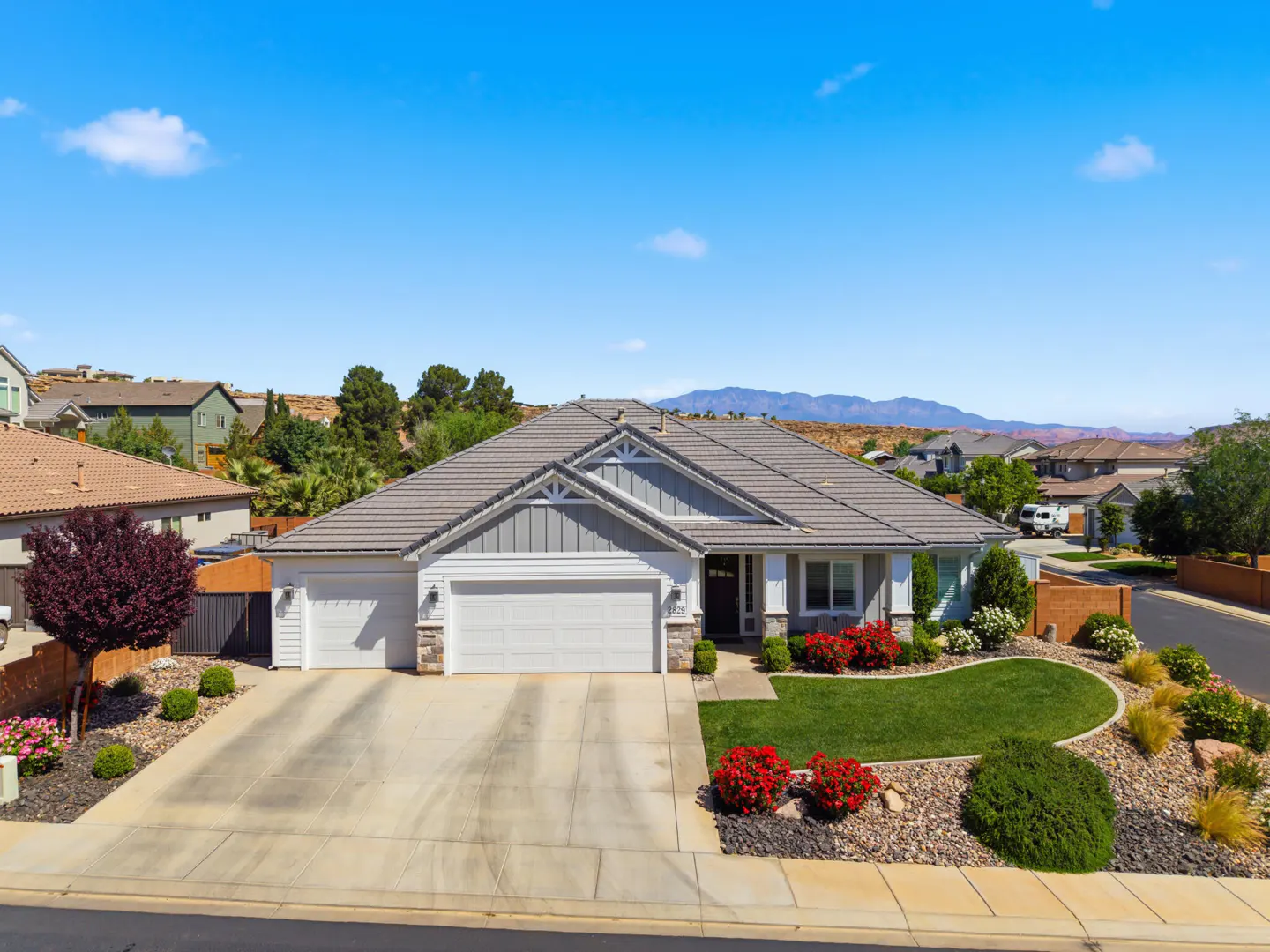 A single-story home with a gray roof, white garage doors, and manicured lawn under a blue sky.