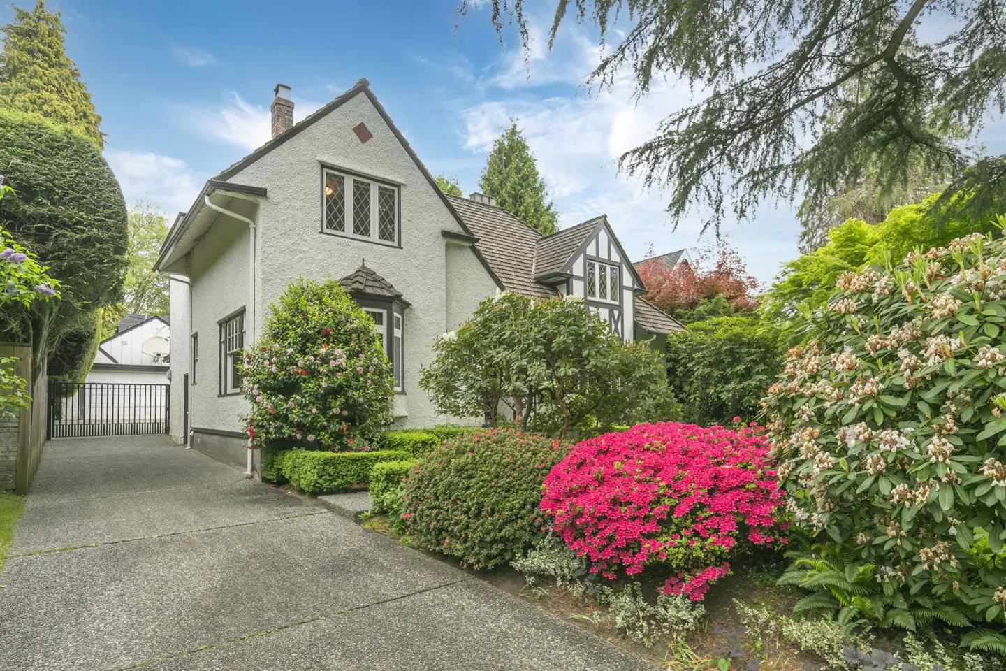 A white Tudor-style house with a brown roof, surrounded by lush green landscaping and bright pink azaleas. A driveway leads to a garage.
