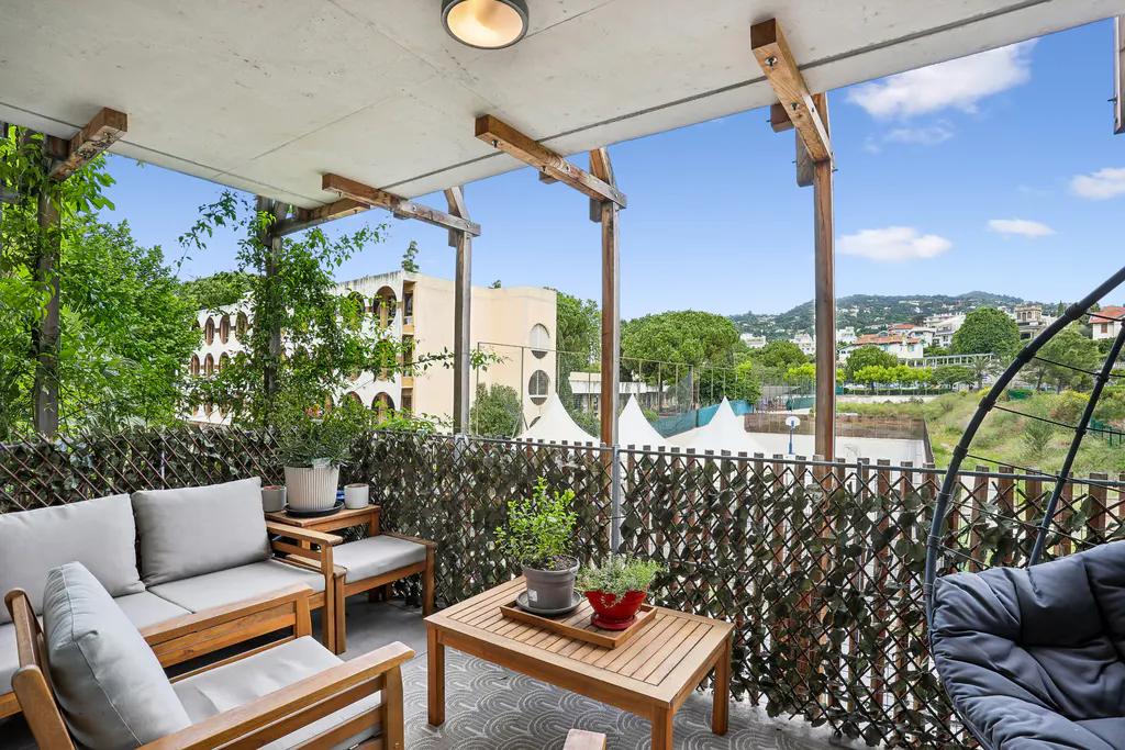 Balcony with wood furniture, gray cushions, and potted plants. A woven screen provides privacy. Views of trees, buildings, and a blue sky.