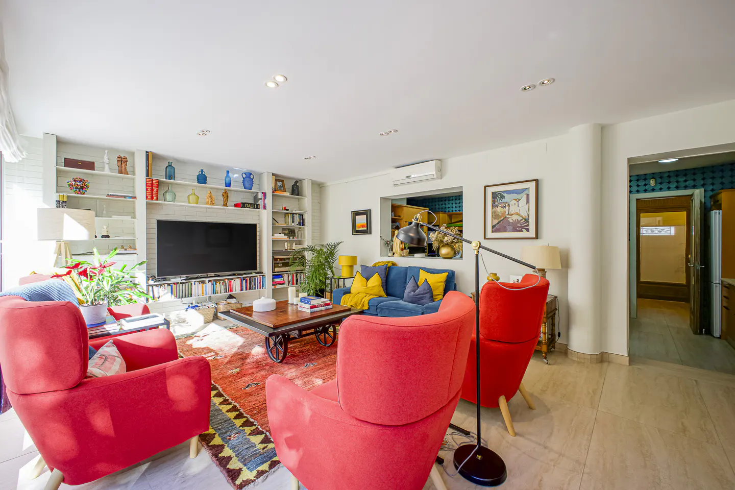 Bright living room with red chairs, blue sofa, and a large bookshelf filled with books and decorative items. A patterned rug adds warmth.