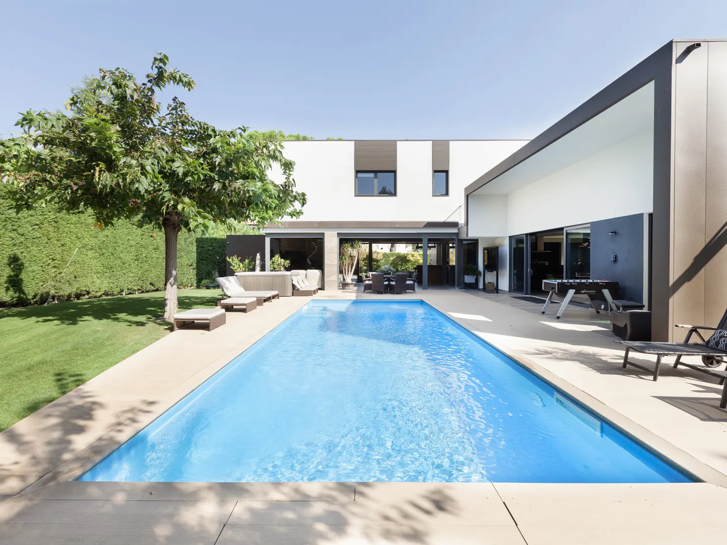 Modern home backyard with a blue swimming pool, lounge chairs, and a foosball table on a sunny day.