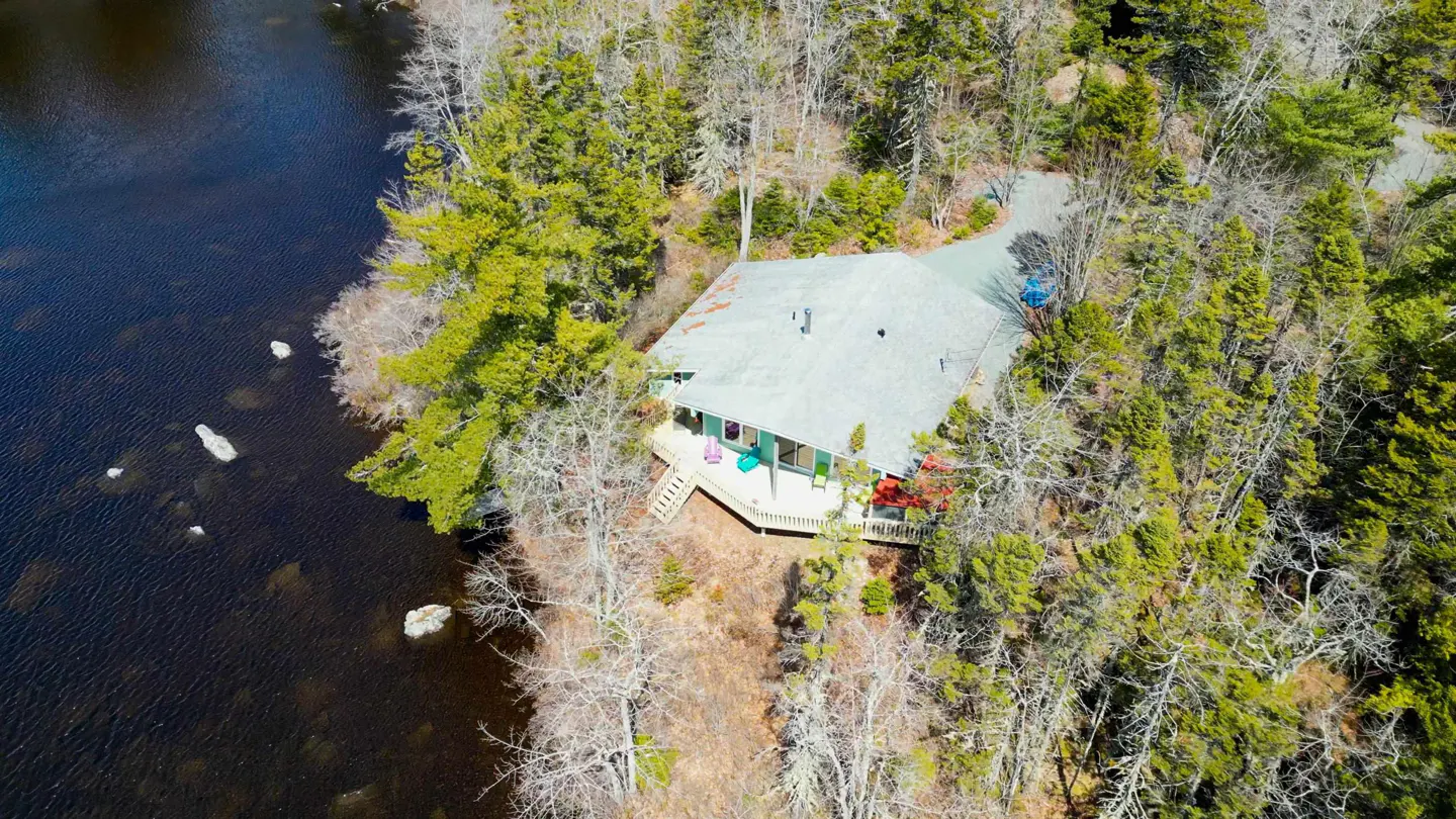 Aerial view of a gray roofed house with a deck, nestled between a dark lake and a forest of green trees.