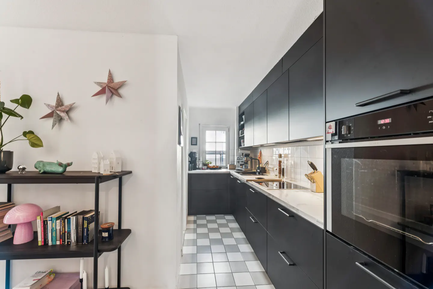 A modern kitchen with black cabinets, white countertops, and a checkered floor. A black shelving unit with books and decor is on the left.