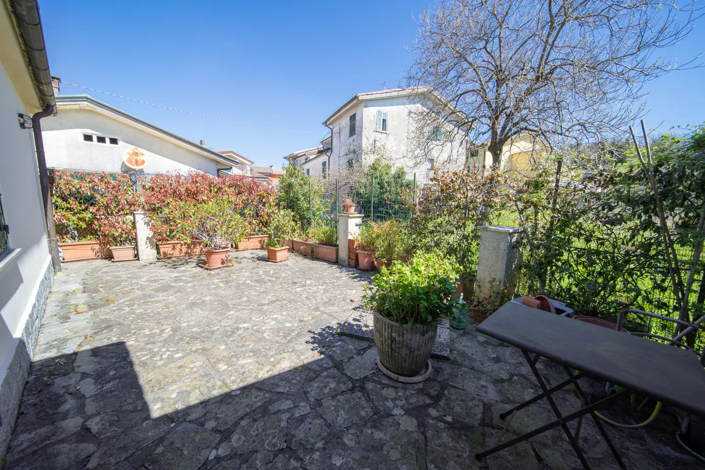Stone patio with potted plants and a small table. A house and trees are in the background under a blue sky.