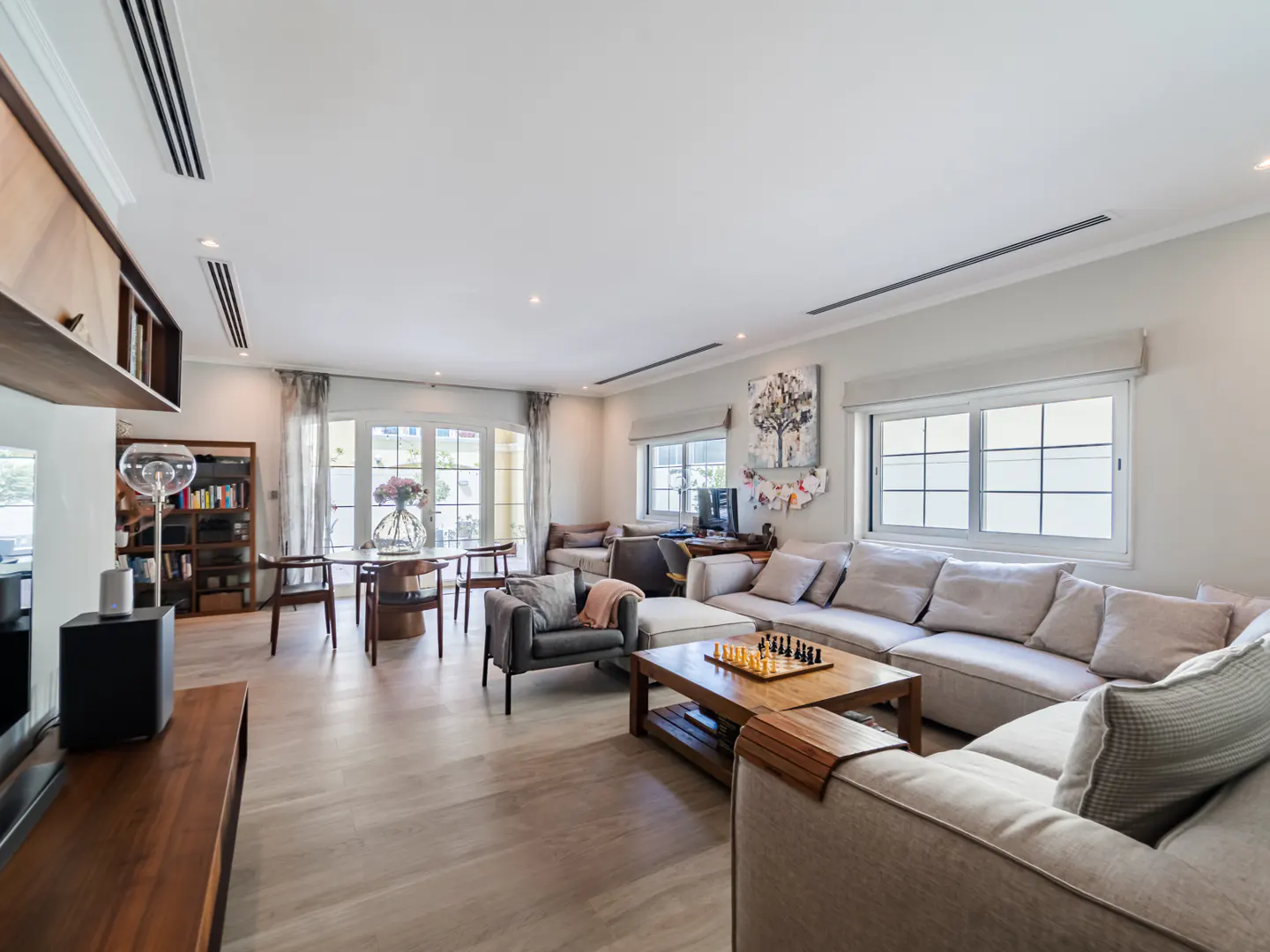 Bright living room with a large beige sectional sofa, a chess board on a wooden coffee table, and a dining table with flowers.