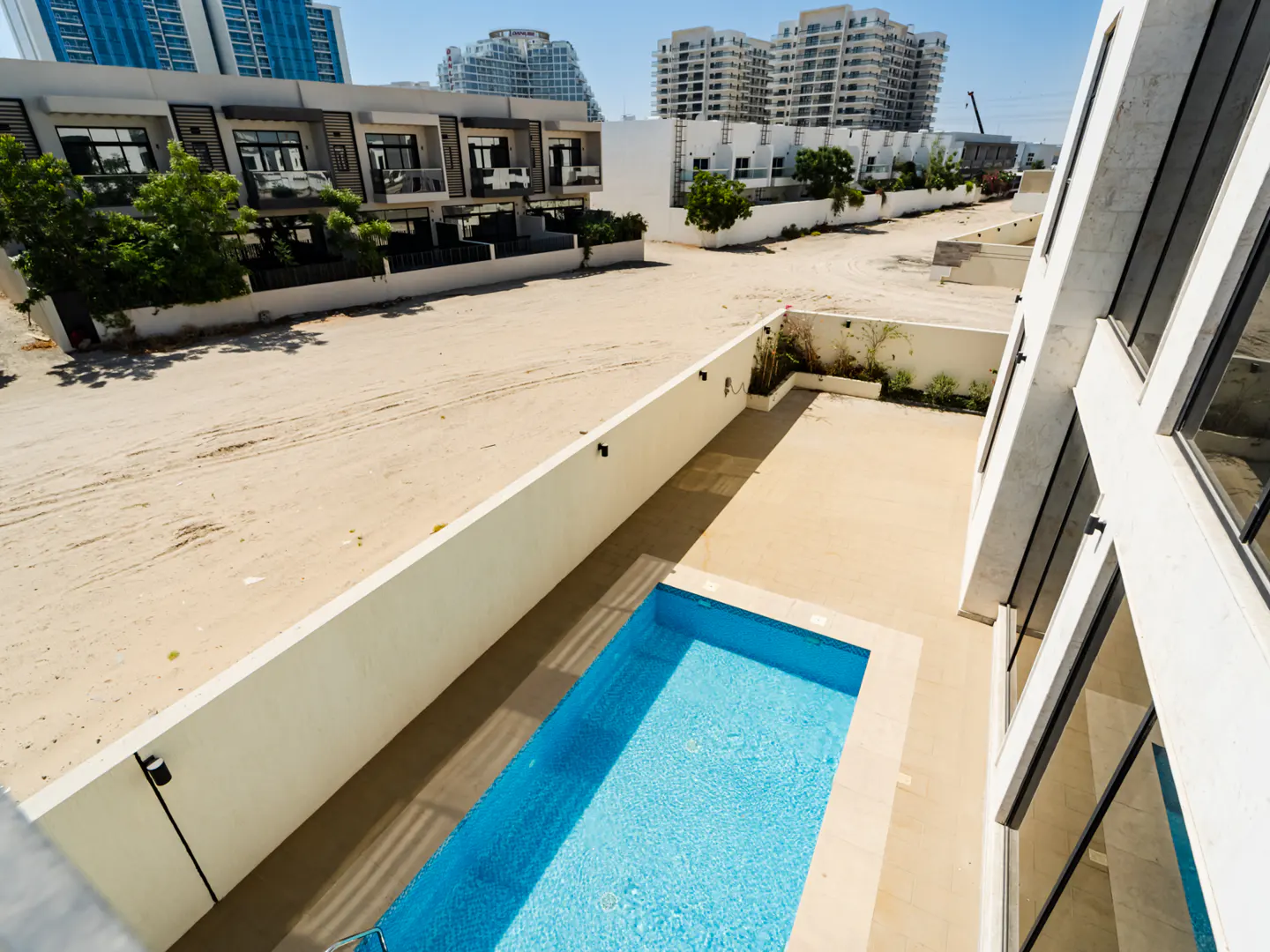 High-angle view of a rectangular blue swimming pool in a modern backyard, with a sandy lot and apartment buildings beyond.