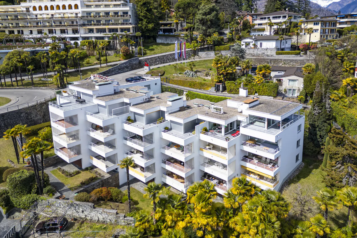 Aerial view of a modern white apartment building with balconies, surrounded by lush green trees and other buildings.