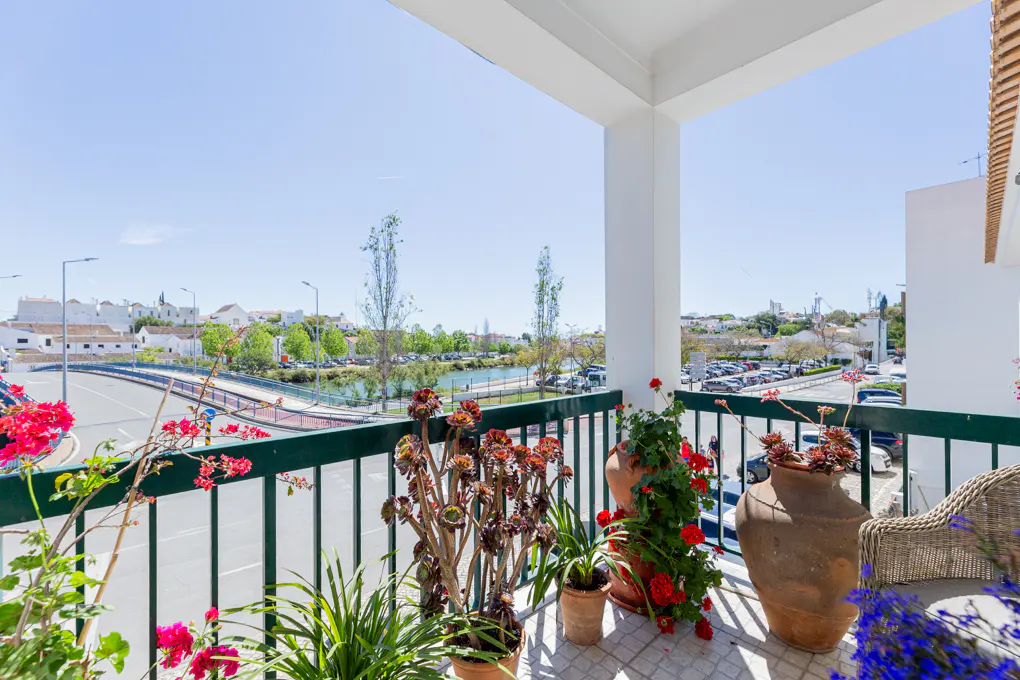 Balcony view with green railing, potted plants, and a wicker chair. A bridge, lake, and white buildings are visible in the background.