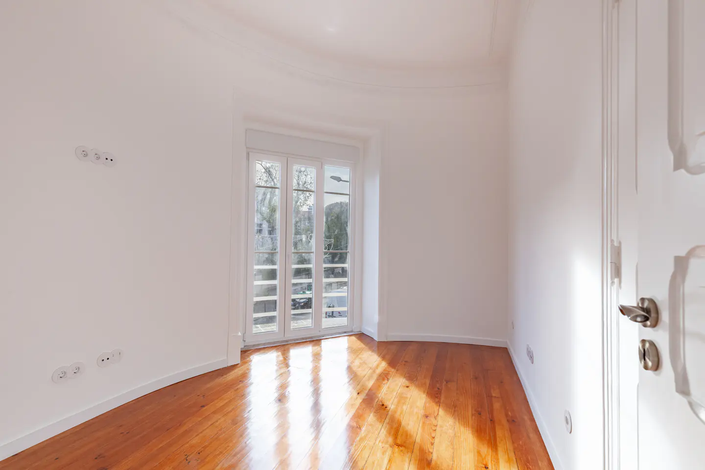Bright, empty room with hardwood floors, white walls, and a multi-pane window showing trees outside. A white door is partially visible.