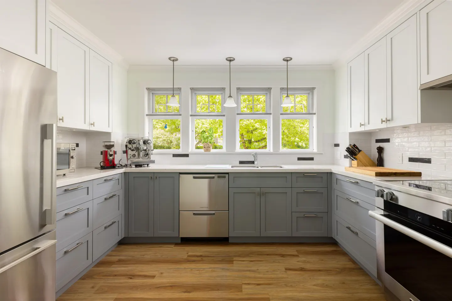 Bright kitchen with gray lower cabinets, white upper cabinets, stainless steel appliances, and wood floors. Three windows overlook green trees.